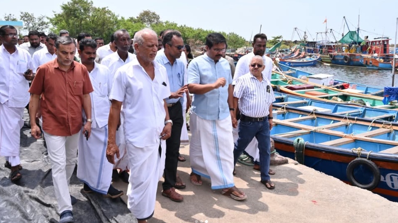 औपचारिक कपड़ों में पुरुषों का एक समूह एक बंदरगाह पर चल रहा है, जहाँ किनारे पर कई मछली पकड़ने वाली नावें खड़ी हैं।