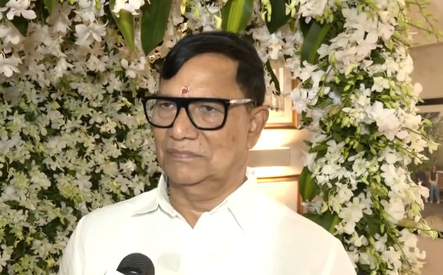 सफेद शर्ट पहने और चश्मा लगाए कृपाशंकर सिंह फूलों की सजावट के सामने खड़े हैं।