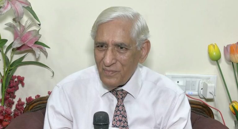 सफेद शर्ट और पैटर्न वाली टाई पहने एक बुजुर्ग व्यक्ति माइक्रोफोन के सामने बैठे हैं।
