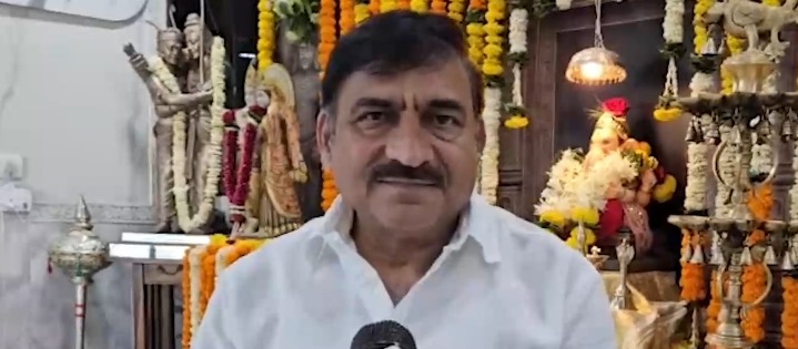 सफेद शर्ट पहने एक व्यक्ति कैमरे की ओर देख रहे हैं, पृष्ठभूमि में धार्मिक मूर्तियां और सजावट है।