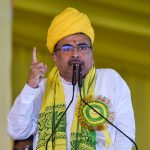 पीली पगड़ी और दुपट्टा पहने एक व्यक्ति मंच पर माइक्रोफोन के सामने भाषण देते हुए अपनी उंगली ऊपर उठाए हुए हैं।