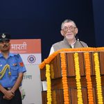 संतोष गंगवार एक मंच पर खड़े होकर भाषण दे रहे हैं, जिसके सामने गेंदे के फूलों की माला लगी है और पास में एक वर्दीधारी सुरक्षाकर्मी खड़ा है।