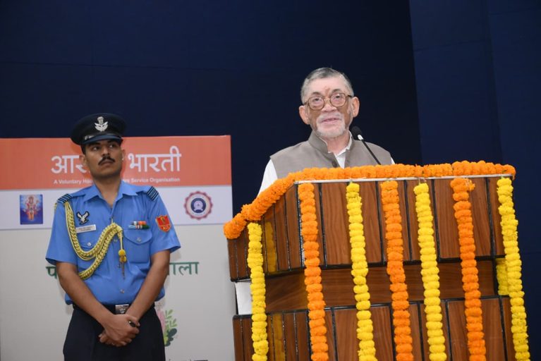 संतोष गंगवार एक मंच पर खड़े होकर भाषण दे रहे हैं, जिसके सामने गेंदे के फूलों की माला लगी है और पास में एक वर्दीधारी सुरक्षाकर्मी खड़ा है।
